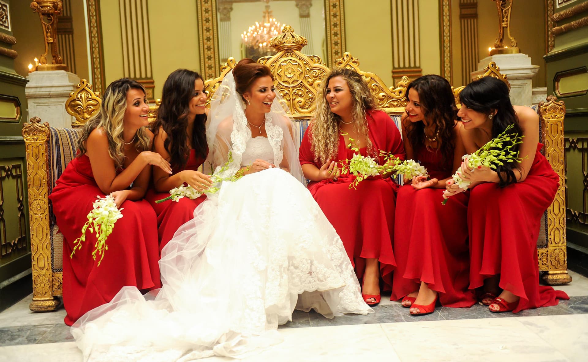 Bride laughing with her bridesmaids in red on a gilded bench