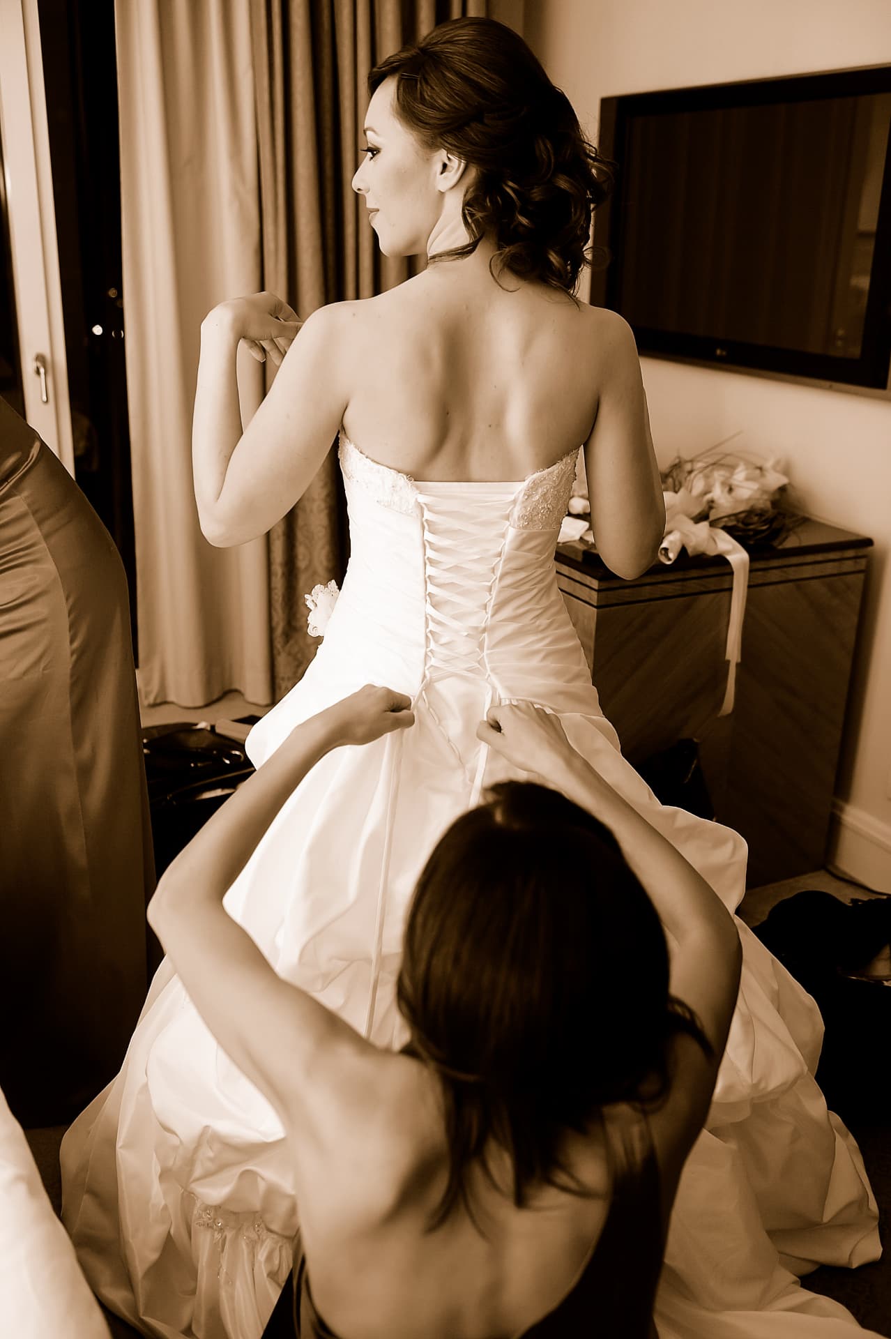 Bridesmaids lacing the bride into her gown in sepia