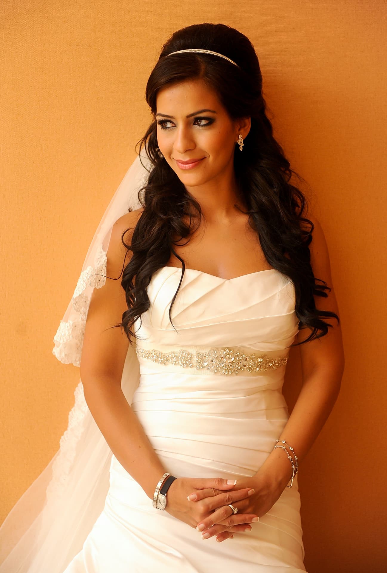 Bride in a satin gown and veil resting against a warm ochre wall