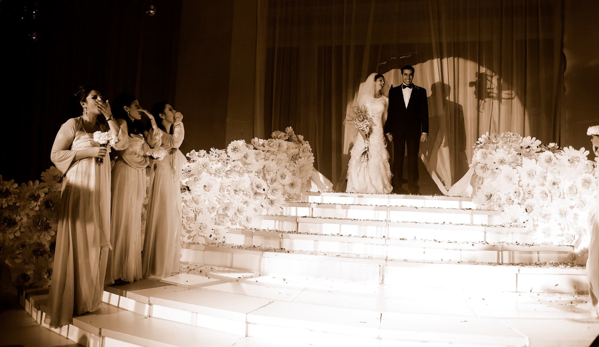 Bride and groom on a lit stage as bridesmaids react with joy